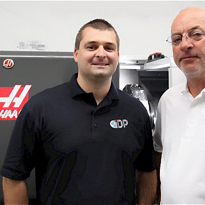 Two IDP team members standing in front of a HAAS CNC machine, both wearing IDP logo shirts and smiling, representing Integrated Defense Products’ leadership.