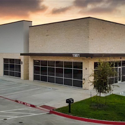 Exterior view of the Integrated Defense Products facility at sunset, showing a modern industrial building with large glass windows, beige brick facade, and an empty parking lot in front.