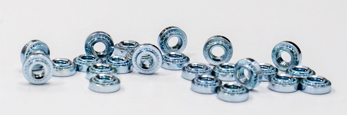 Close-up view of various fasteners, including self-clinching, broaching, surface mount, and weld-on types, arranged on a metallic surface to showcase different shapes and finishes.